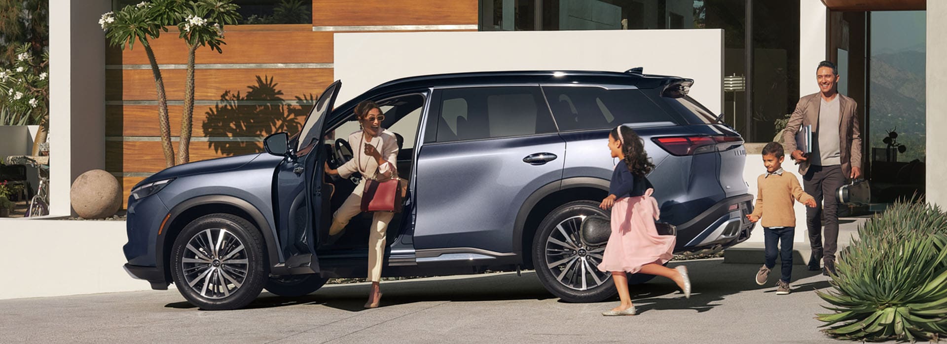 Woman getting out of driver seat of INFINITI QX60 being greeted by her two young children and husband