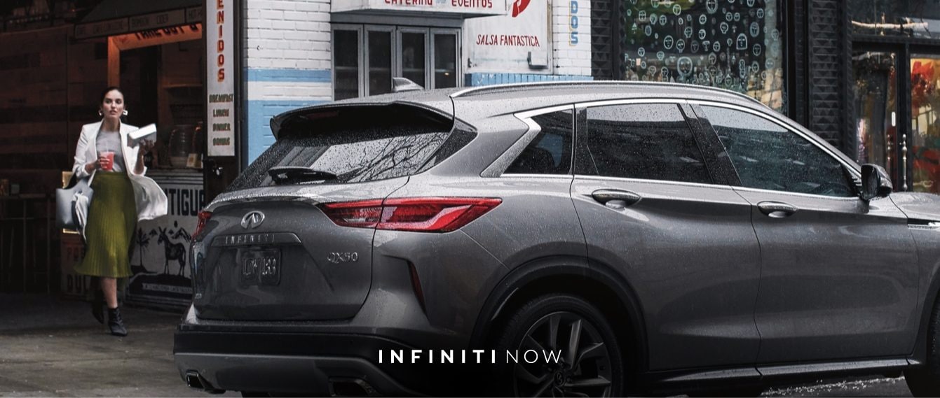 INFINITI Now | Woman walking towards 2020 INFINITI QX50 luxury crossover parked on street shown in graphite shadow color