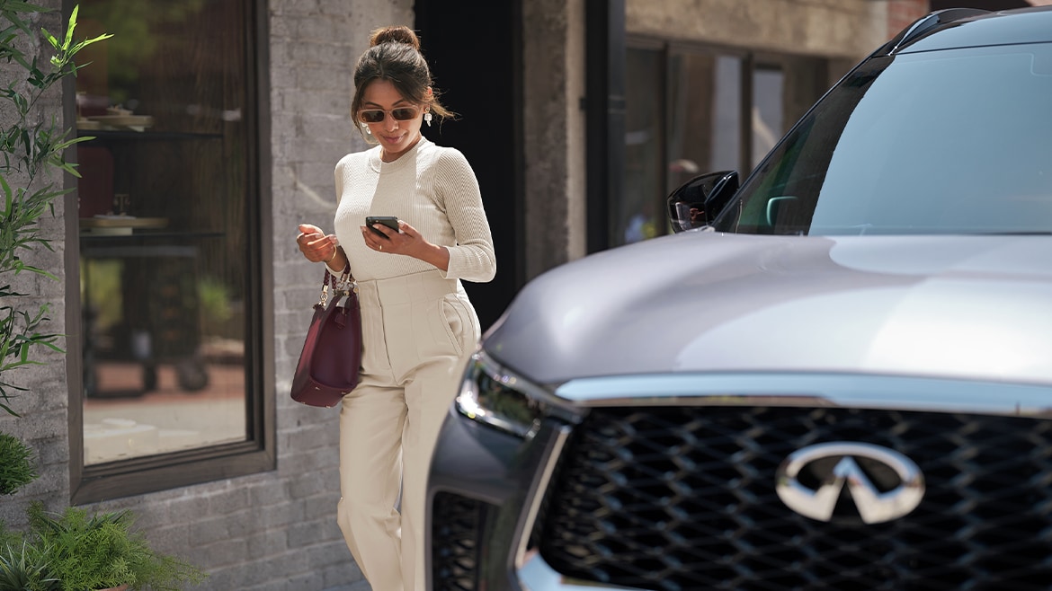 Woman looking at her phone while walking beside the front exterior of a 2022 INFINITI QX60 Crossover SUV