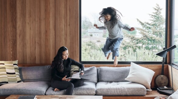 Woman sitting and reading book on a white couch while young girl jumps off couch