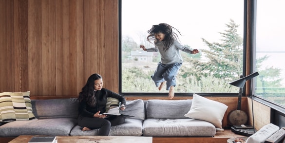 Woman sitting on couch and reading a book while daughter jumps on couch